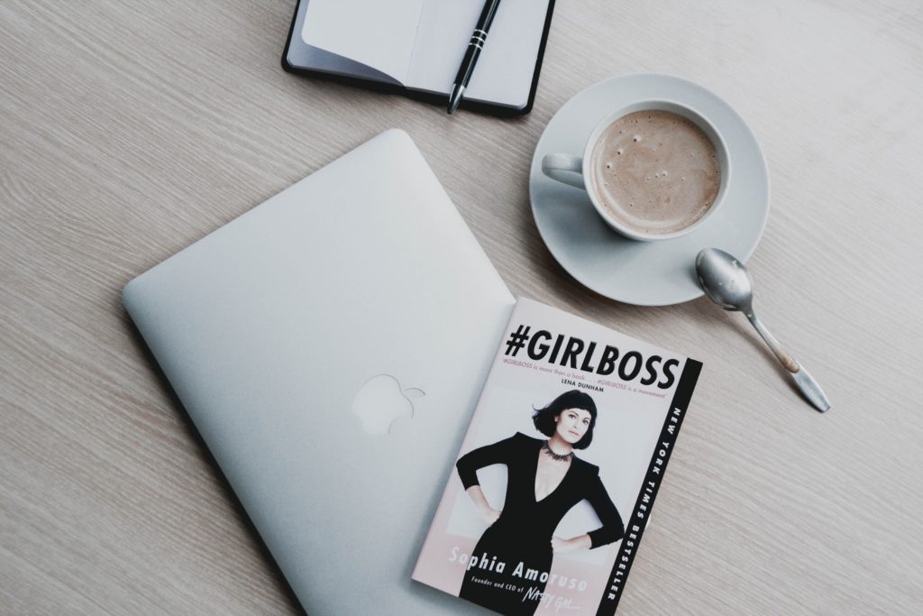 A layflat picture of an Macbook sitting on a table with a magazine titled "Girl Boss" and a cup of coffee. 