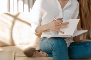 A picture of a woman in a white shirt and jeans sitting on a tan couch opening a notebook to write in it.