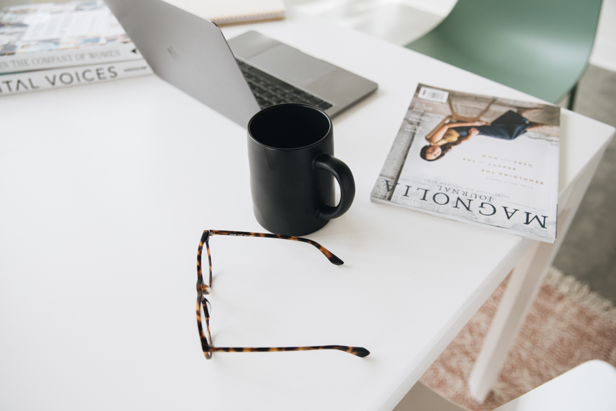 A detailed picture of an open laptop, coffee cup, magazine, and pair of glasses sitting on a white desk. 
