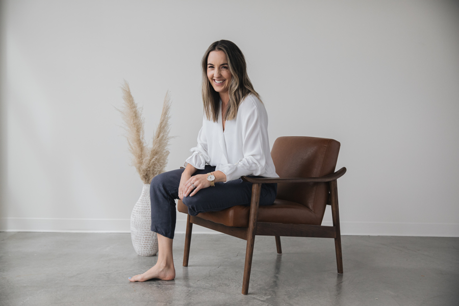 A candid picture of a woman in a white top and black pants sitting cross legged in a brown leather chair in front of a white wall. 