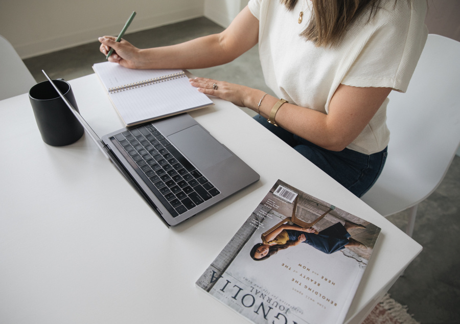 A cropped picture of a woman in a white shirt sitting at a desk writing on a notepad, a laptop, coffee cup, and magazine sitting on the table in front of her. 