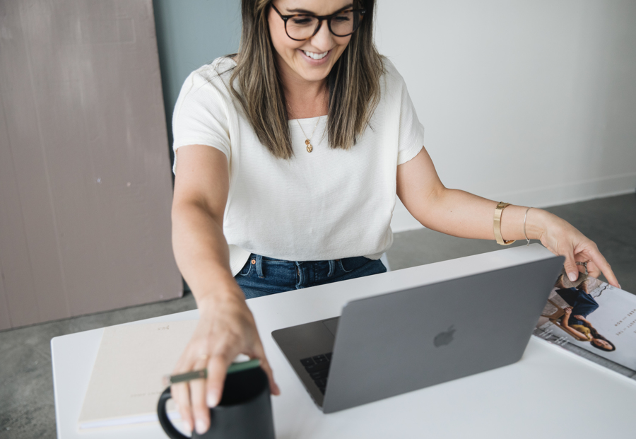 A candid picture of a woman in a white shirt sitting at a desk with a laptop in front of her, reaching for a coffee mug with a smile on her face. 
